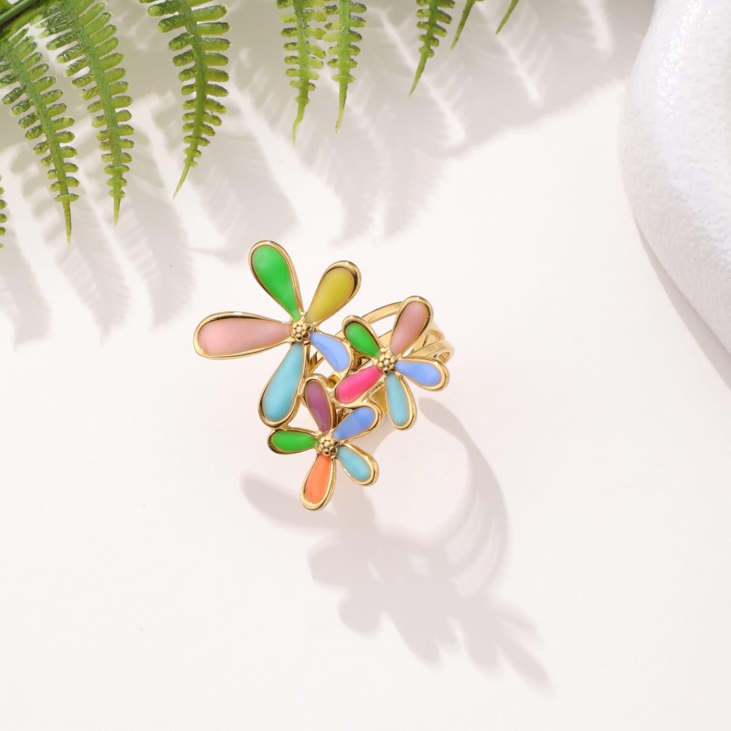 Bague fantaisie avec trois fleurs aux pétales colorés (vert, rose, bleu, orange, jaune) bordés de doré, posée sur une surface claire avec des feuilles de fougère en arrière-plan.