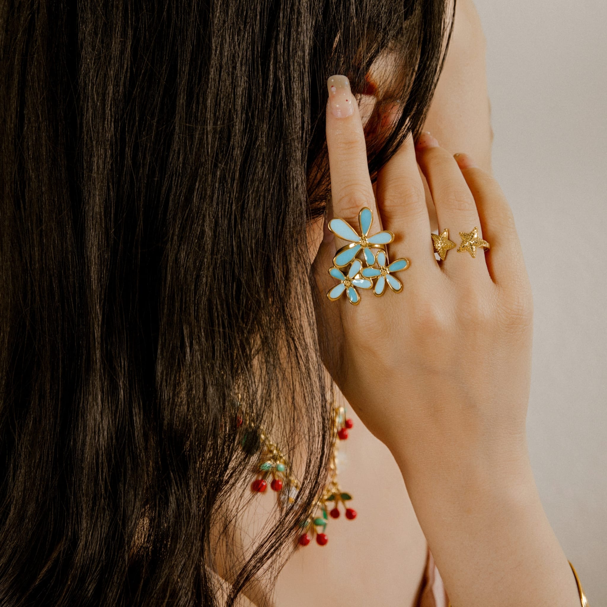 Main d’une personne portant deux bagues : une grande bague florale bleue et dorée, et une bague dorée en forme d’étoiles, accompagnées d’un collier doré avec perles rouges et vertes.