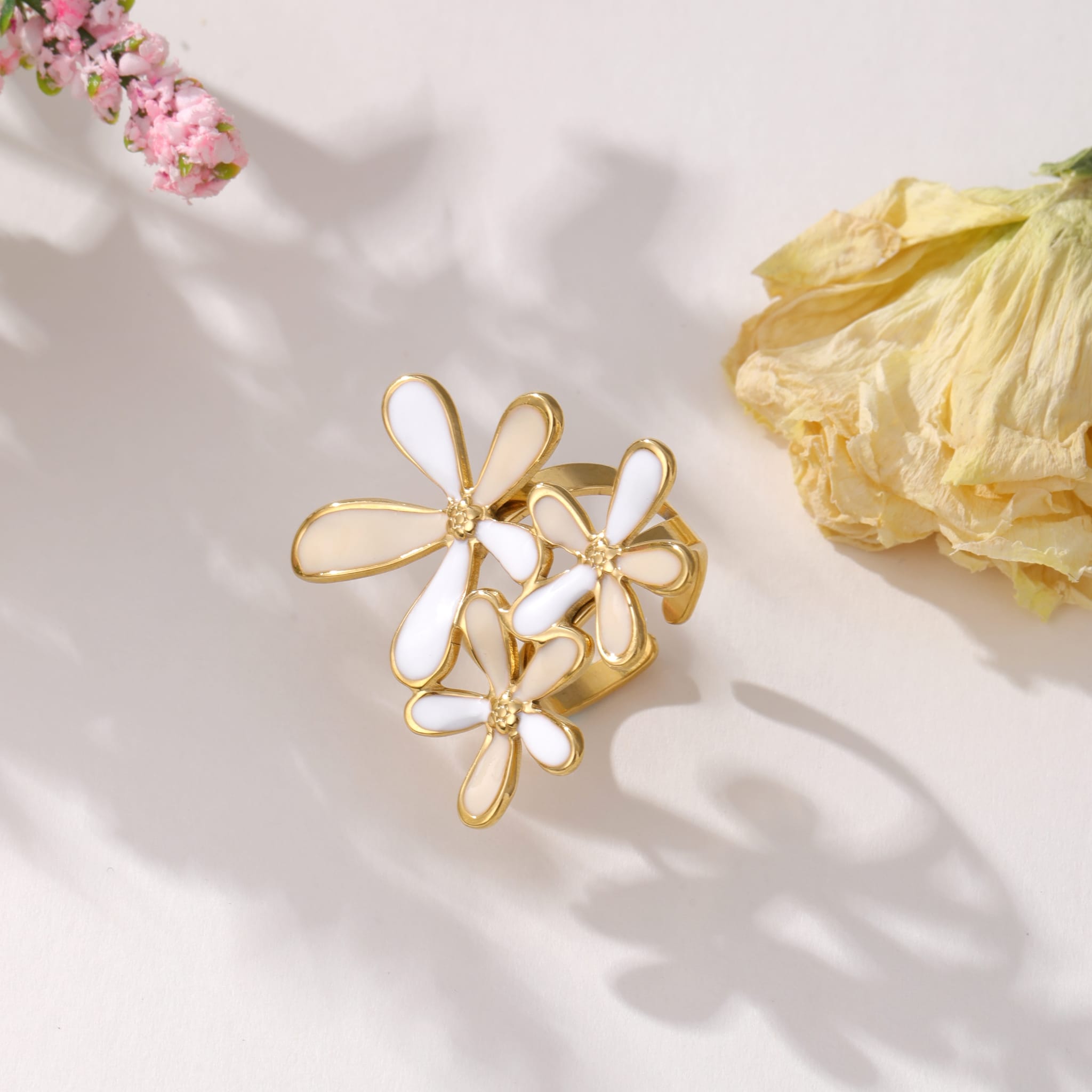 Bague dorée avec trois fleurs en émail blanc, posée sur une surface claire avec des ombres florales et deux fleurs séchées en arrière-plan.