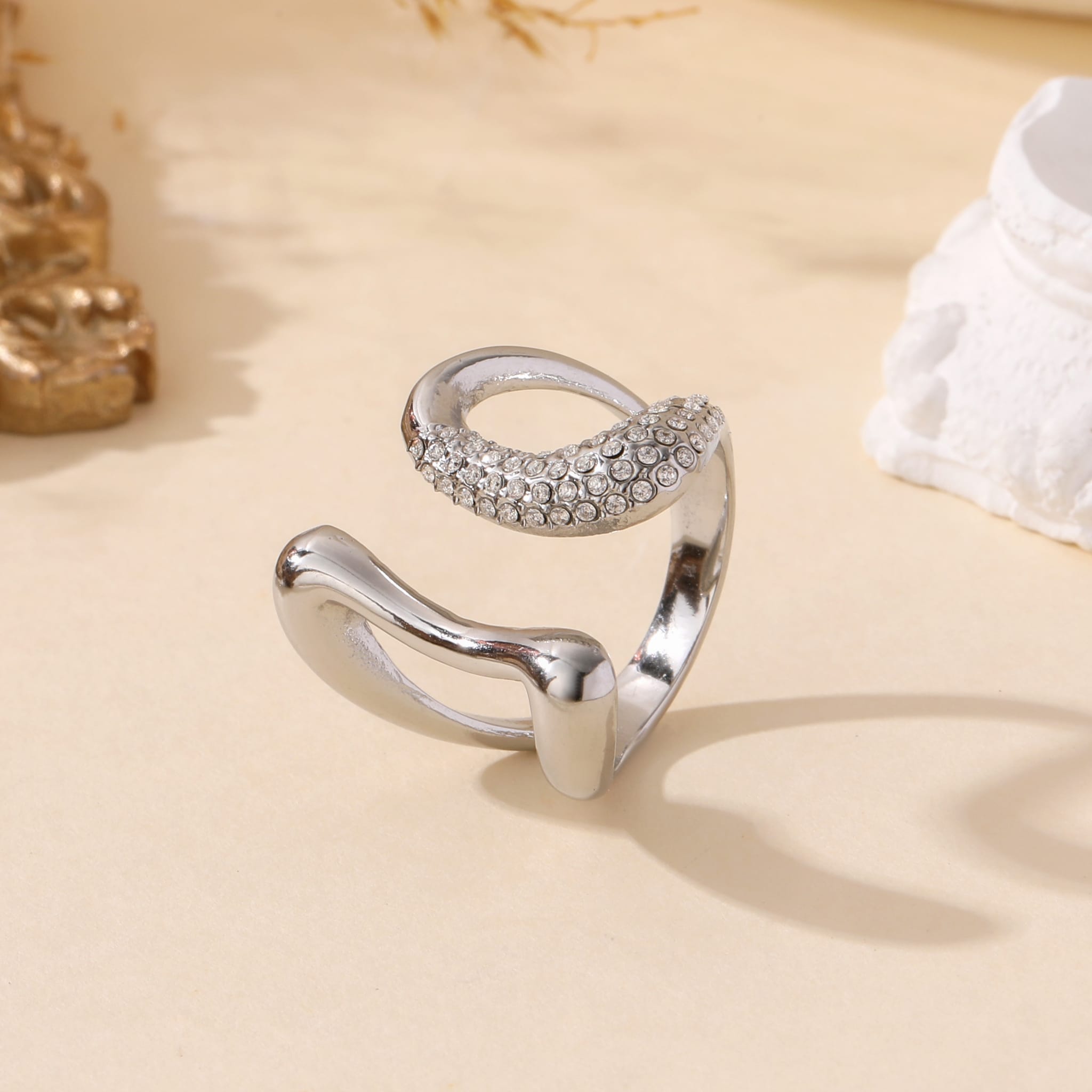Bague ouverte en acier inoxydable argenté, sertie de strass étincelants, au style élégant et contemporain, posée sur fond beige