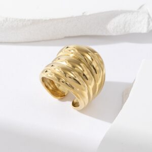 Bague Rava en acier inoxydable doré, au design texturé et ondulé, posée sur fond blanc avec fragments décoratifs en relief.