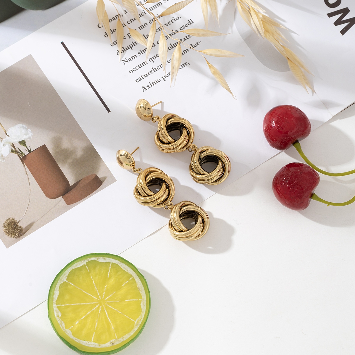 Boucles d’oreilles Naya en acier inoxydable doré, au design torsadé et entrelacé, présentées sur fond blanc avec des cerises, une tranche de citron vert, des tiges de blé et une page imprimée minimaliste.