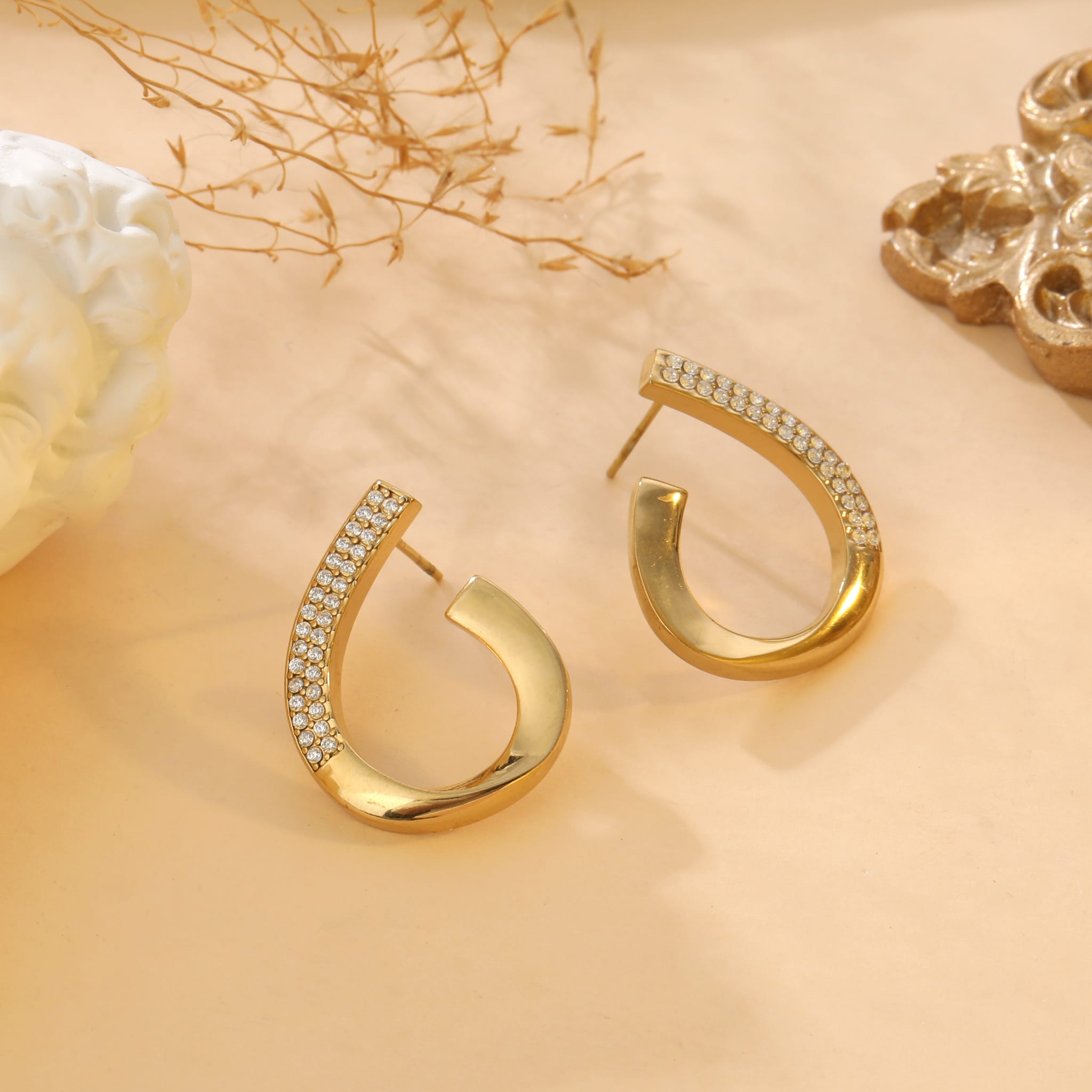 Boucles d’oreilles Alyra en acier inoxydable doré, en forme de goutte sertie de pierres scintillantes, présentées sur fond beige avec des éléments décoratifs naturels et dorés.
