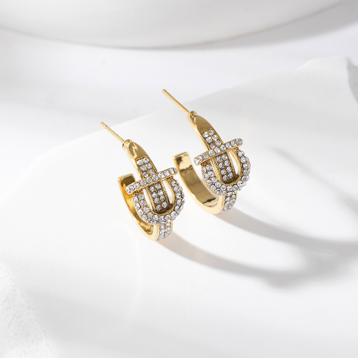 Boucles d’oreilles Diora en acier inoxydable doré, ornées de pierres scintillantes et d’un motif inspiré d’une boucle de ceinture, présentées sur fond blanc avec une lumière douce qui met en valeur leur brillance et leur design audacieux.