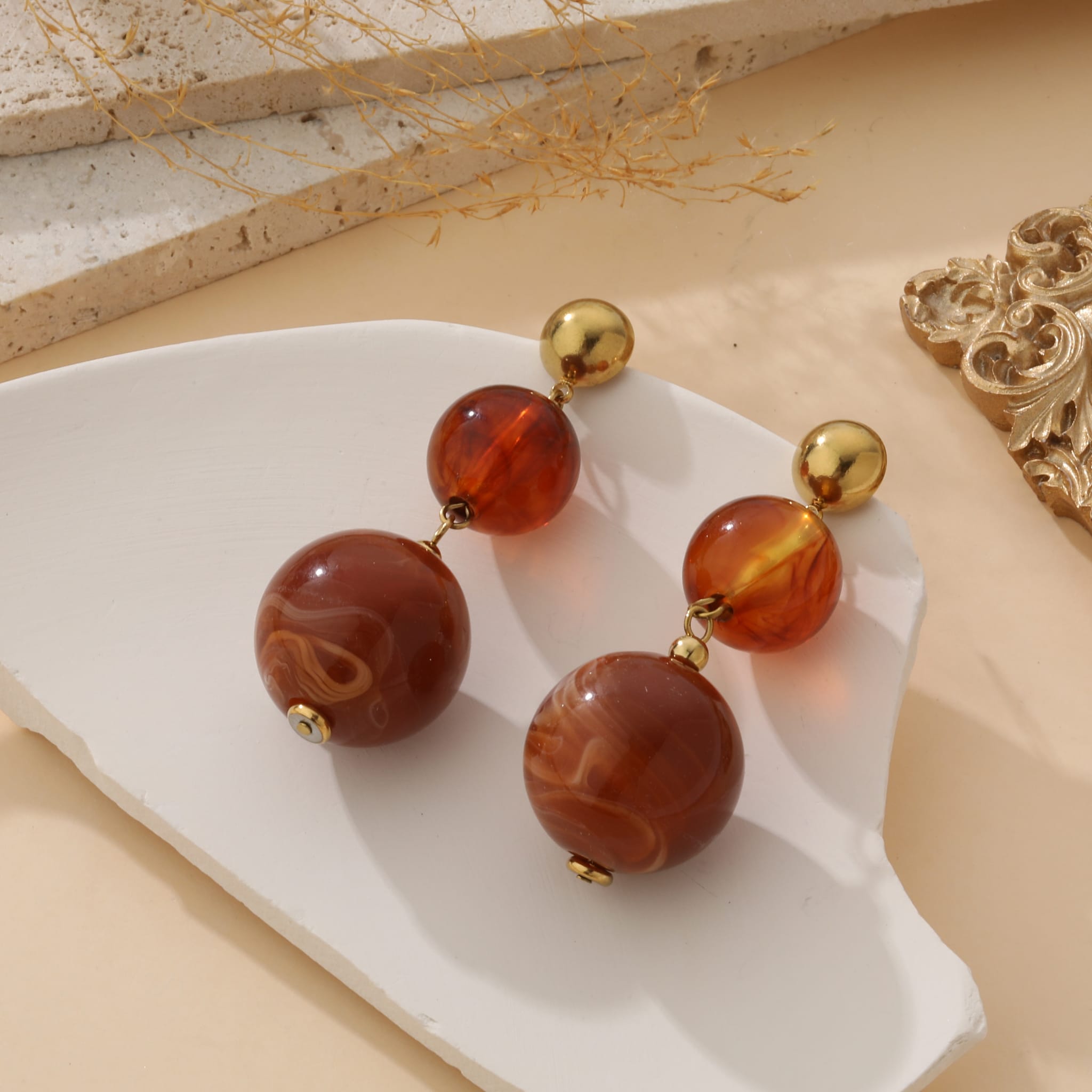 Boucles d’oreilles fantaisie en acier inoxydable doré, ornées de perles marbrées crème et beige, présentées sur fond blanc avec éléments décoratifs naturels – bijou élégant et lumineux pour femme.