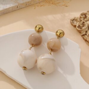 Boucles d’oreilles fantaisie en acier inoxydable doré, ornées de perles marbrées crème et beige, présentées sur fond blanc avec éléments décoratifs naturels – bijou élégant et lumineux pour femme.