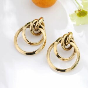 Boucles d'oreilles en acier inoxydable, anneaux entrelacés dorés, présentées sur fond blanc avec mandarine et fleur jaune pâle – bijou élégant et intemporel pour femme.