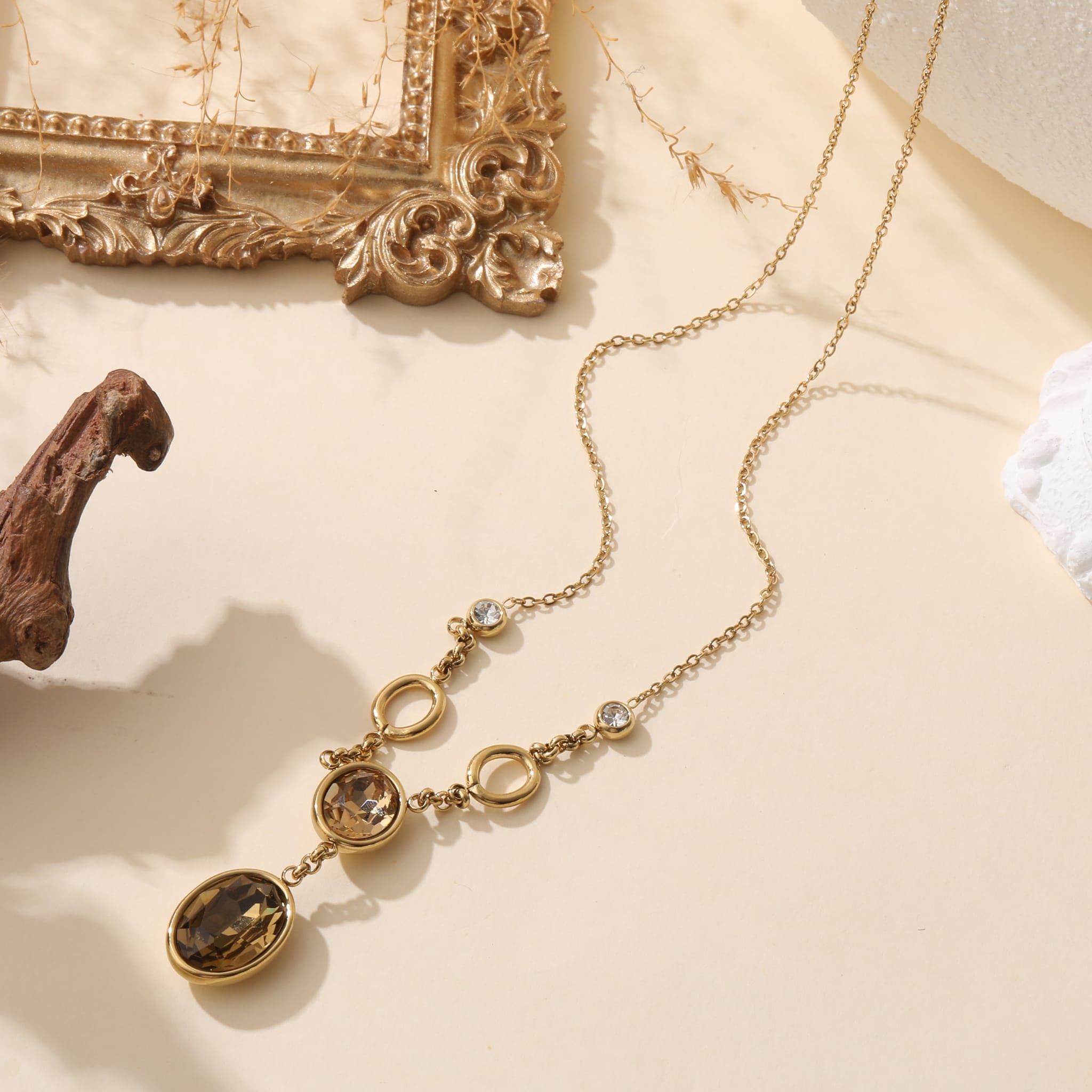 Collier fantaisie en acier inoxydable avec pendeloques Cristaux sur chaîne en maillon fin, présenté sur fond beige avec cadre doré et décor organique – bijou élégant et intemporel pour femme.