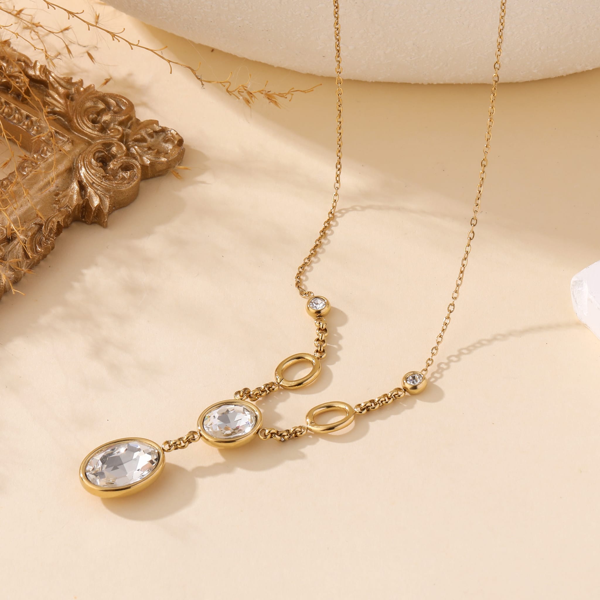 Collier fantaisie en acier inoxydable avec pendeloques Cristaux sur chaîne en maillon fin, présenté sur fond beige avec cadre doré et décor organique – bijou élégant et intemporel pour femme.