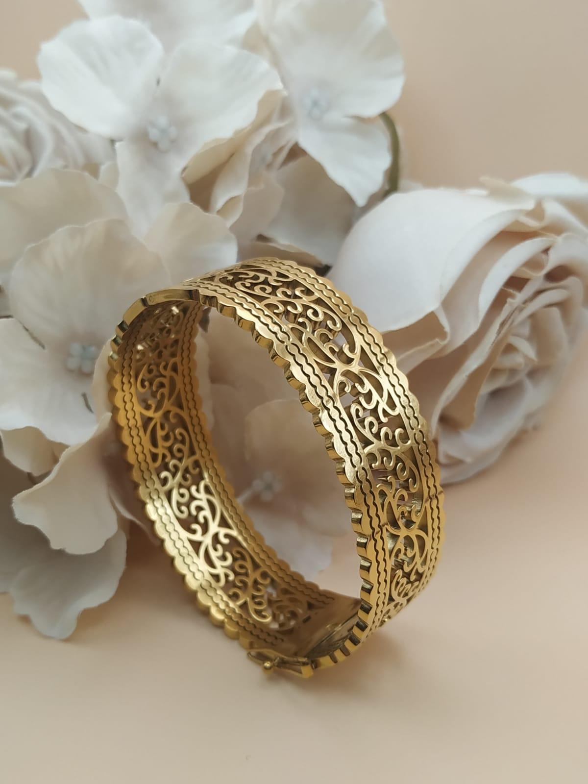 Bracelet jonc doré au motif ajouré de style oriental, posé devant des roses blanches et beige sur fond doux.