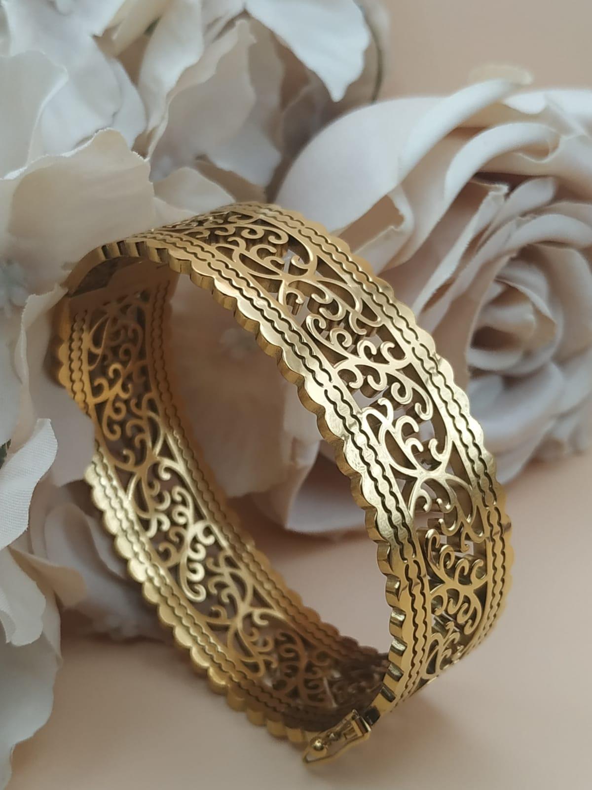 Bracelet jonc doré au motif ajouré de style oriental, posé devant des roses blanches et beige sur fond doux.