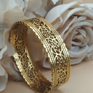 Bracelet jonc doré au motif ajouré de style oriental, posé devant des roses blanches et beige sur fond doux.