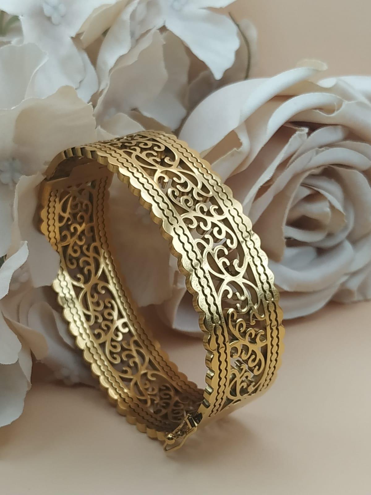 Bracelet jonc doré au motif ajouré de style oriental, posé devant des roses blanches et beige sur fond doux.