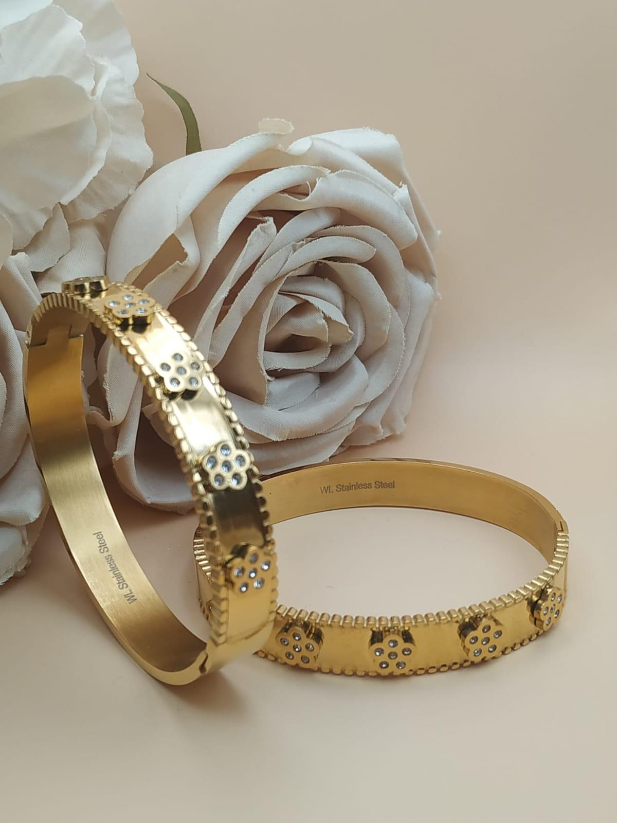 Bracelet doré rigide avec motifs floraux sertis de pierres claires, posé devant des roses blanches et roses sur fond beige.