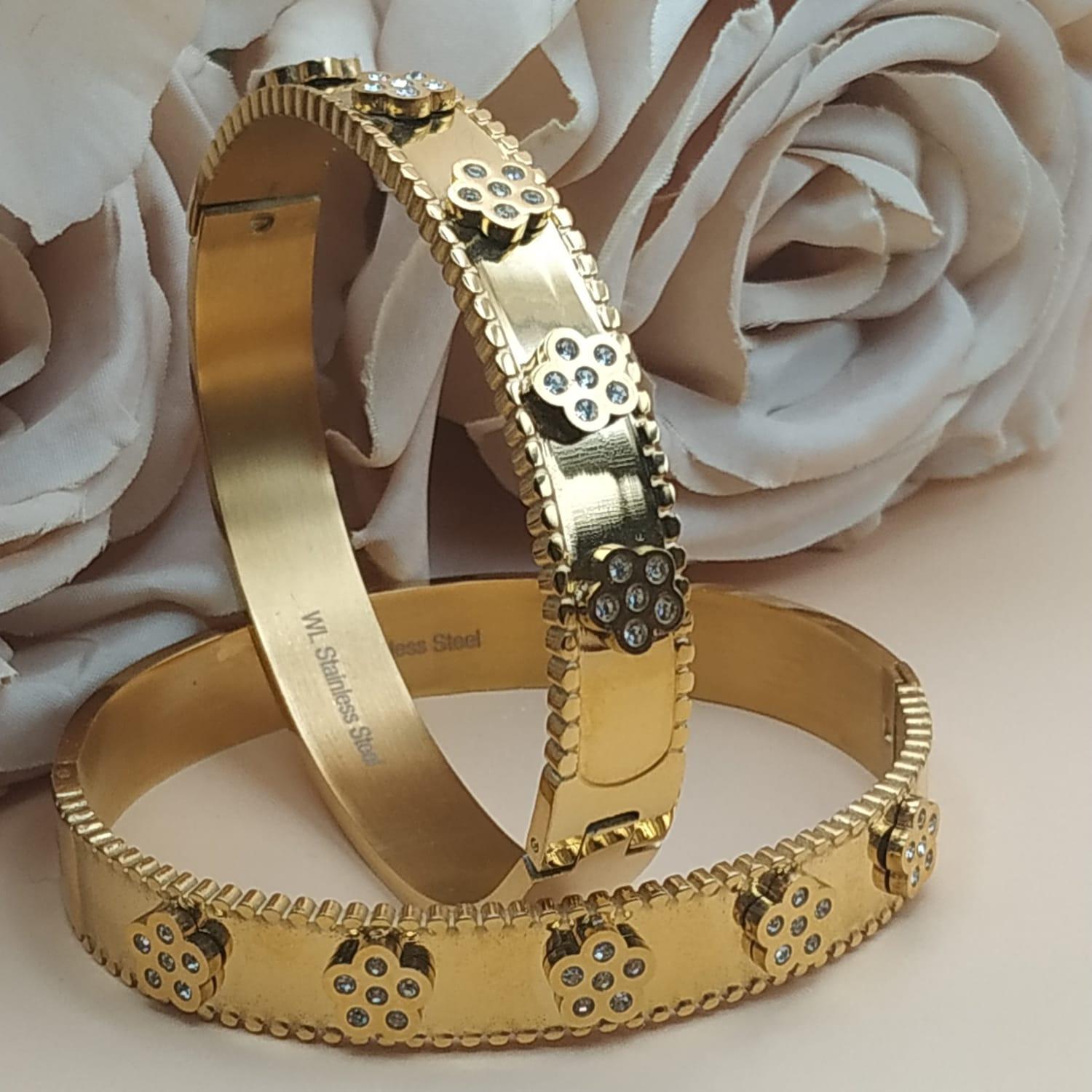 Bracelet doré rigide avec motifs floraux sertis de pierres claires, posé devant des roses blanches et roses sur fond beige.