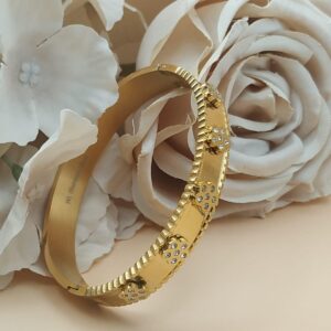 Bracelet doré rigide avec motifs floraux sertis de pierres claires, posé devant des roses blanches et roses sur fond beige.