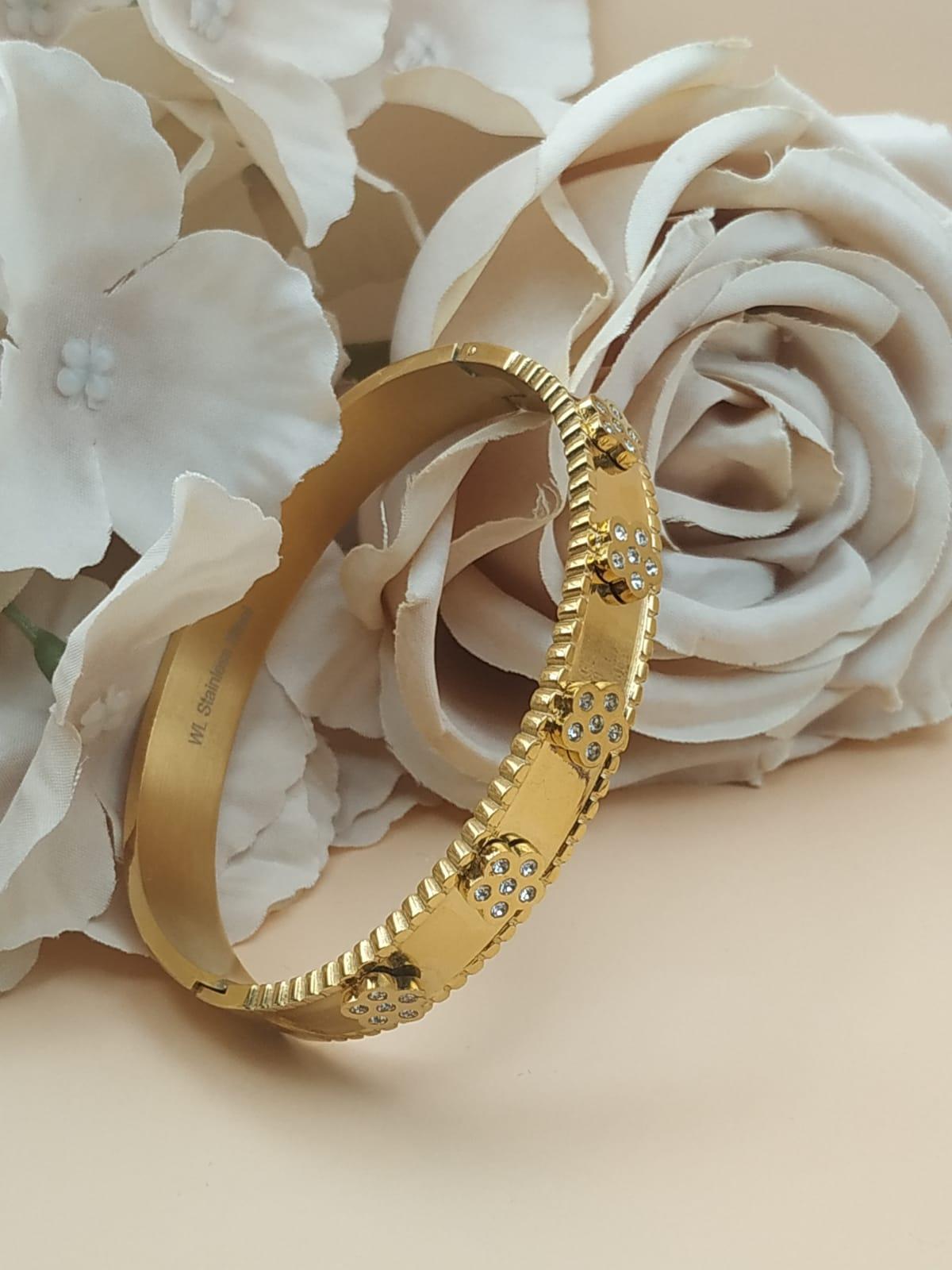 Bracelet doré rigide avec motifs floraux sertis de pierres claires, posé devant des roses blanches et roses sur fond beige.