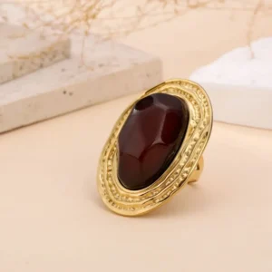 Bague dorée avec grand cabochon ovale rouge, sertie dans un anneau texturé au style vintage, posée sur fond clair avec éléments naturels.