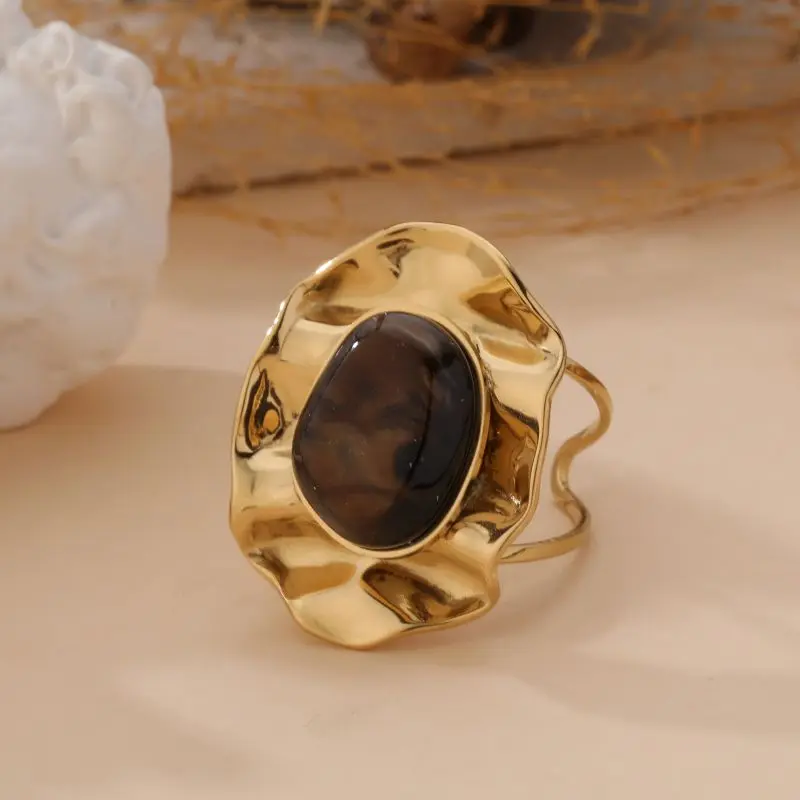 Bague dorée à forme ondulée avec cabochon brun marbré, sertie dans un anneau organique au style artisanal, posée sur fond beige avec éléments naturels.