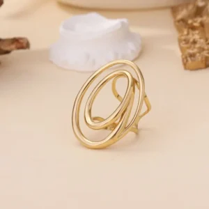 Bague dorée composée de deux anneaux ovales entrelacés, au design abstrait et moderne, posée sur fond beige avec éléments décoratifs.