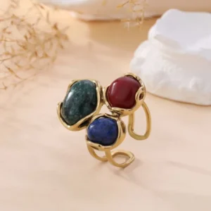 Bague en or à double anneau ornée de trois pierres serties : une verte marbrée, une rouge profonde et une bleue mouchetée. Les pierres sont disposées en composition artistique sur fond neutre avec éléments végétaux séchés.