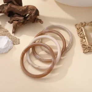 Bracelets jonc Augustine en résine brillante, disponibles en plusieurs couleurs naturelles, posés sur fond beige avec éléments décoratifs.