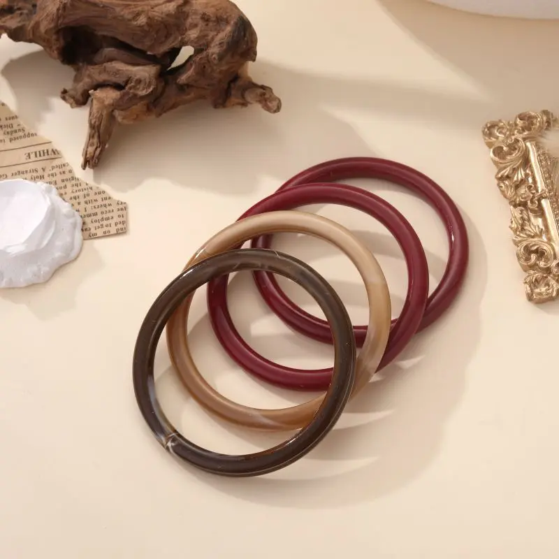 Bracelets jonc Augustine en résine brillante, disponibles en plusieurs couleurs naturelles, posés sur fond beige avec éléments décoratifs.