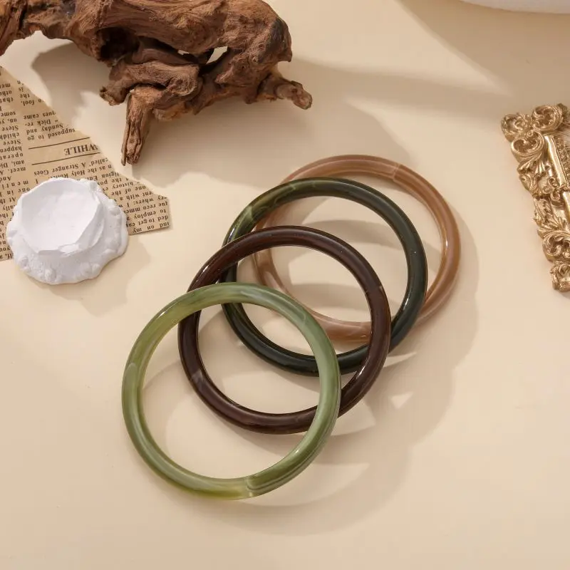 Bracelets jonc Augustine en résine brillante, disponibles en plusieurs couleurs naturelles, posés sur fond beige avec éléments décoratifs.