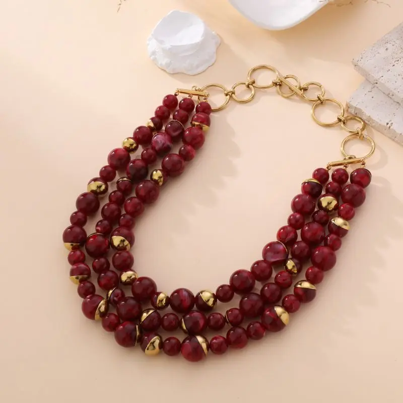 Collier multi-rangs Laura avec perles rouges et dorées, fermoir en anneaux dorés, bijou audacieux présenté sur fond beige