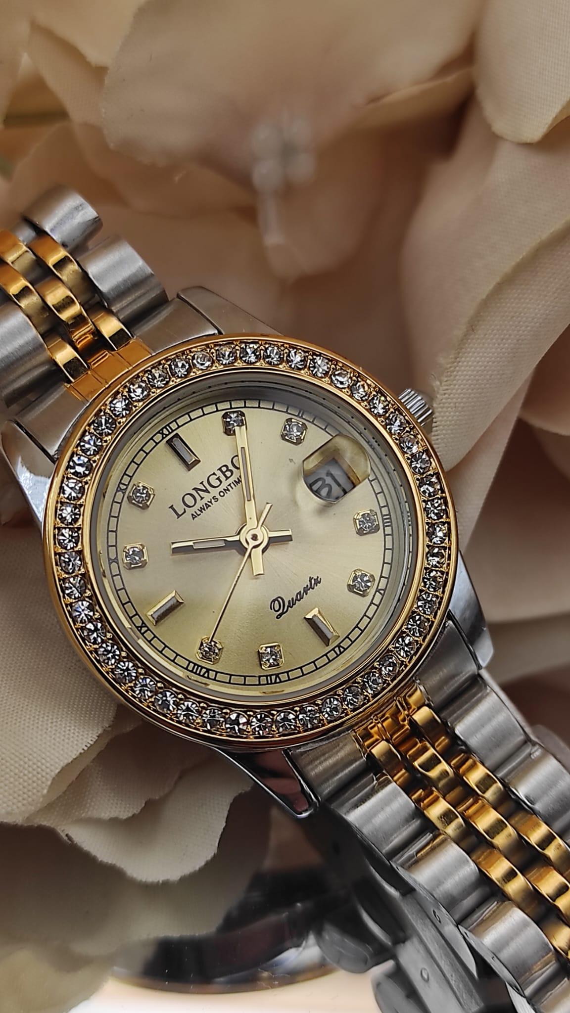 Montre LONGBO à cadran doré avec bracelet bicolore argent et or, lunette sertie de cristaux, date avec loupe à 3h, posée sur un fond de fleurs beige