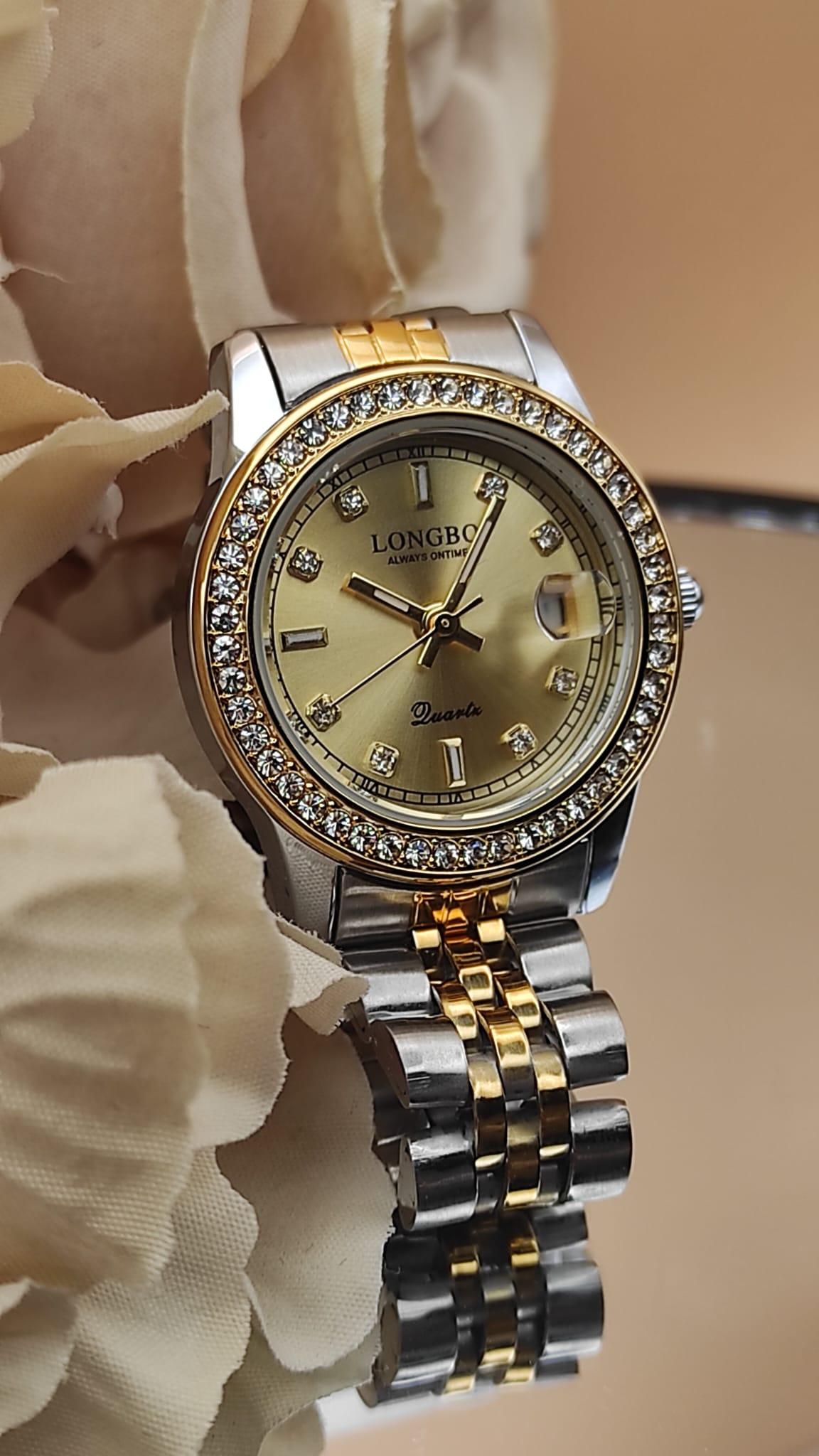 Montre LONGBO à cadran doré avec bracelet bicolore argent et or, lunette sertie de cristaux, date avec loupe à 3h, posée sur un fond de fleurs beige