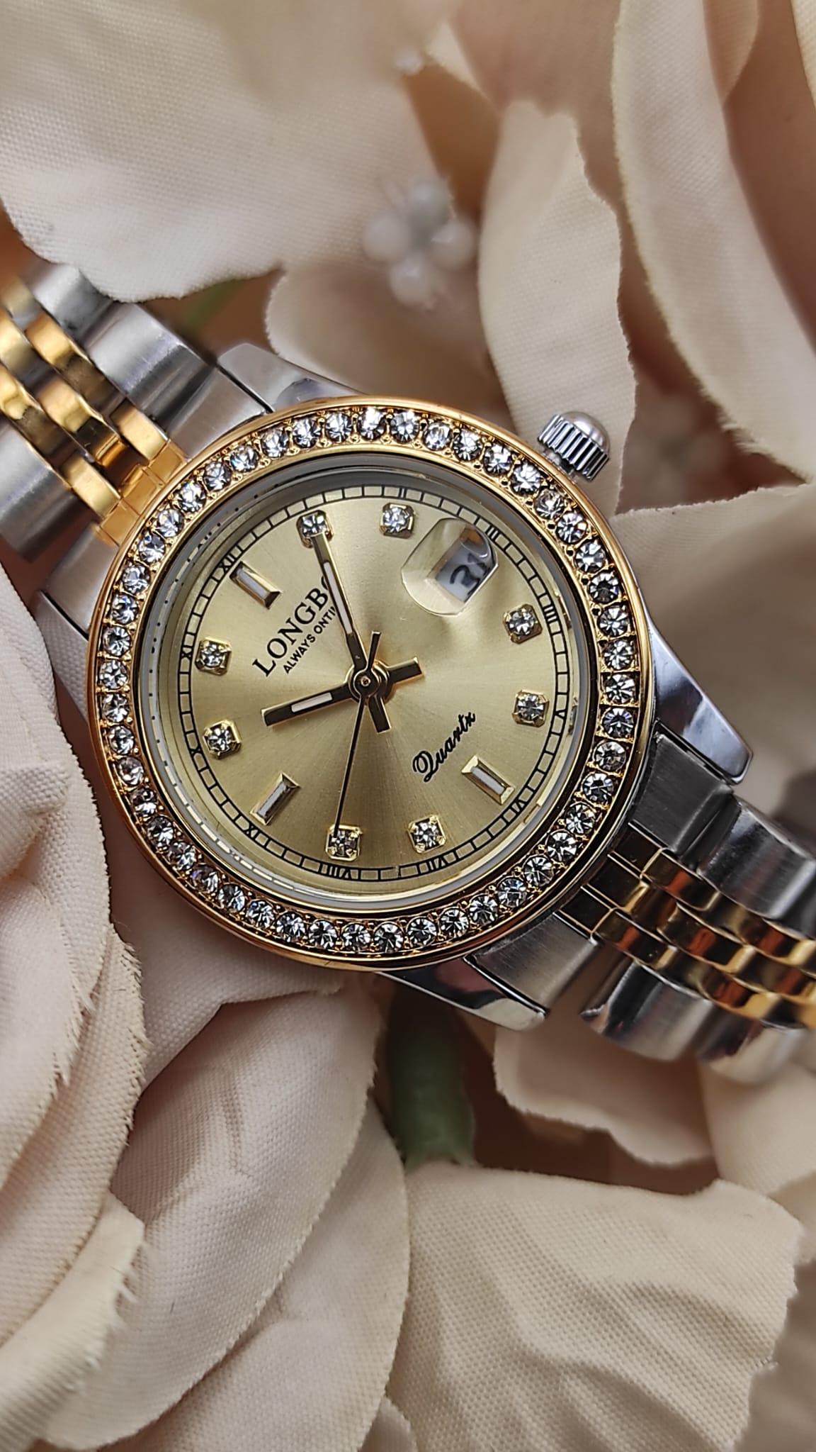 Montre LONGBO à cadran doré avec bracelet bicolore argent et or, lunette sertie de cristaux, date avec loupe à 3h, posée sur un fond de fleurs beige