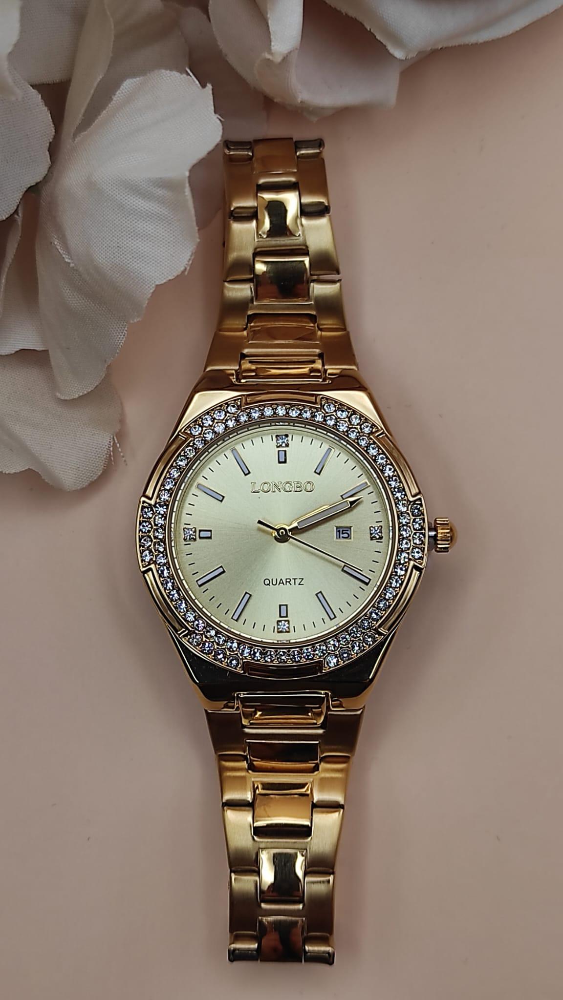 Montre dorée LONGBO avec cadran doré, lunette sertie de cristaux brillants et bracelet métallique, posée sur un fond de pétales beige.