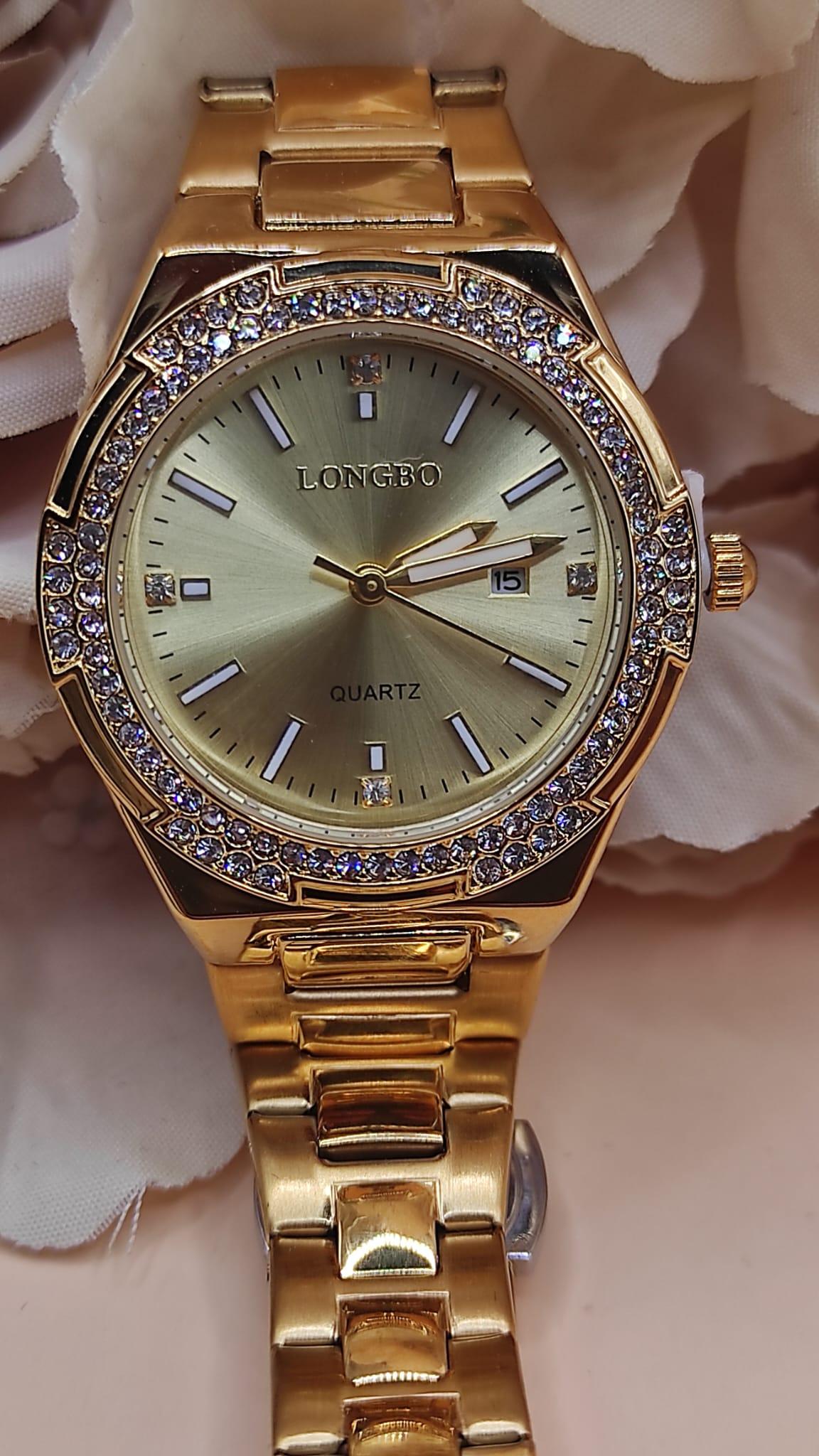 Montre dorée LONGBO avec cadran doré, lunette sertie de cristaux brillants et bracelet métallique, posée sur un fond de pétales beige.