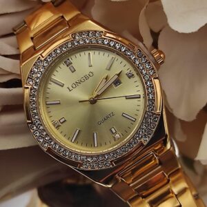 Montre dorée LONGBO avec cadran doré, lunette sertie de cristaux brillants et bracelet métallique, posée sur un fond de pétales beige.