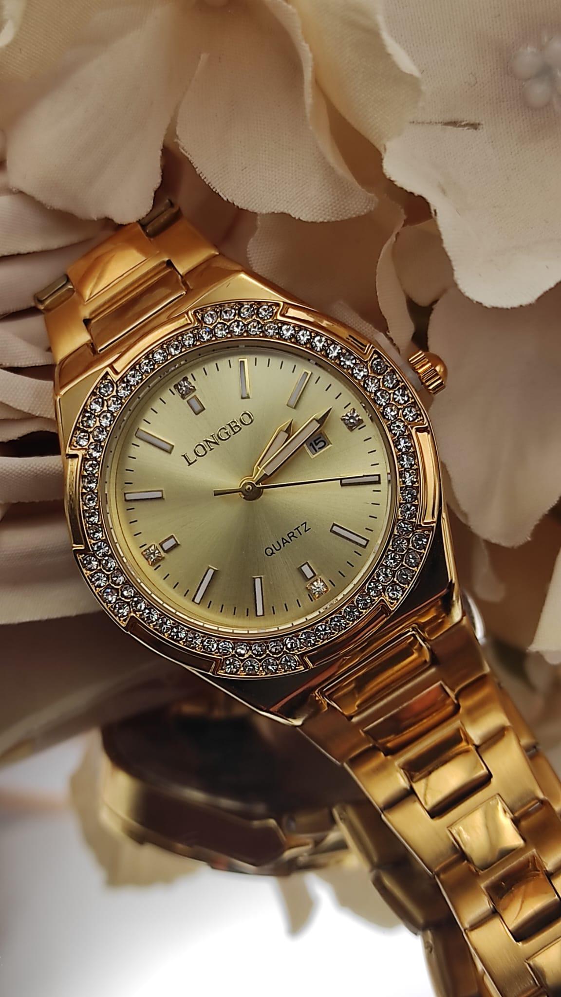 Montre dorée LONGBO avec cadran doré, lunette sertie de cristaux brillants et bracelet métallique, posée sur un fond de pétales beige.