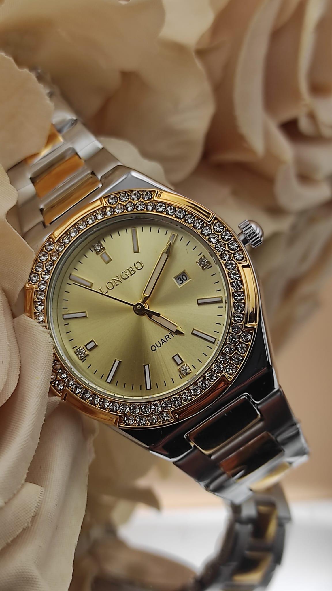 Montre LONGBO à cadran doré avec bracelet bicolore argent et or, lunette sertie de cristaux brillants, posée sur un fond de fleurs beige.