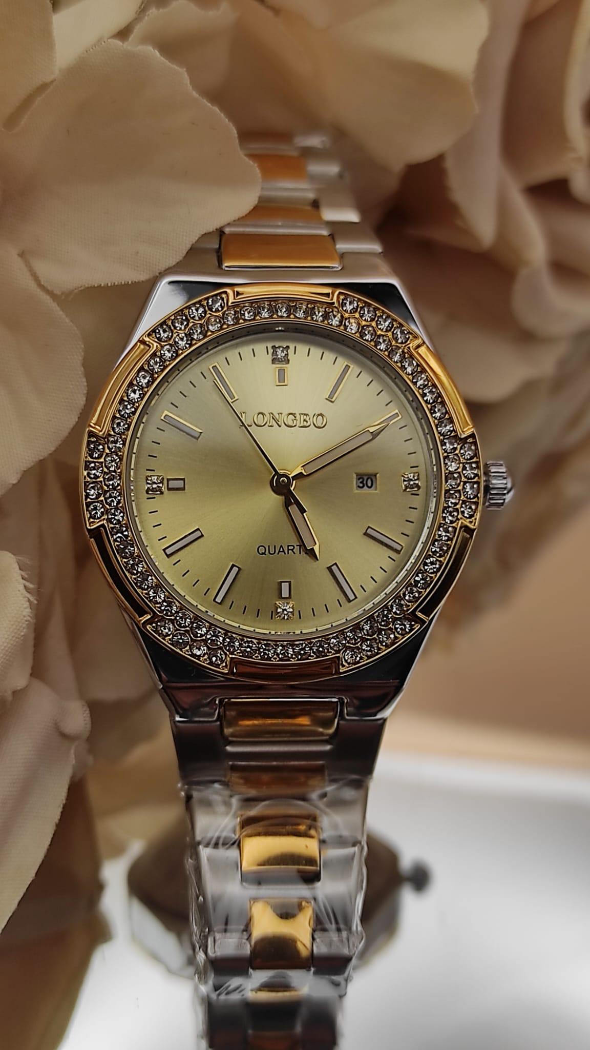 Montre LONGBO à cadran doré avec bracelet bicolore argent et or, lunette sertie de cristaux brillants, posée sur un fond de fleurs beige.