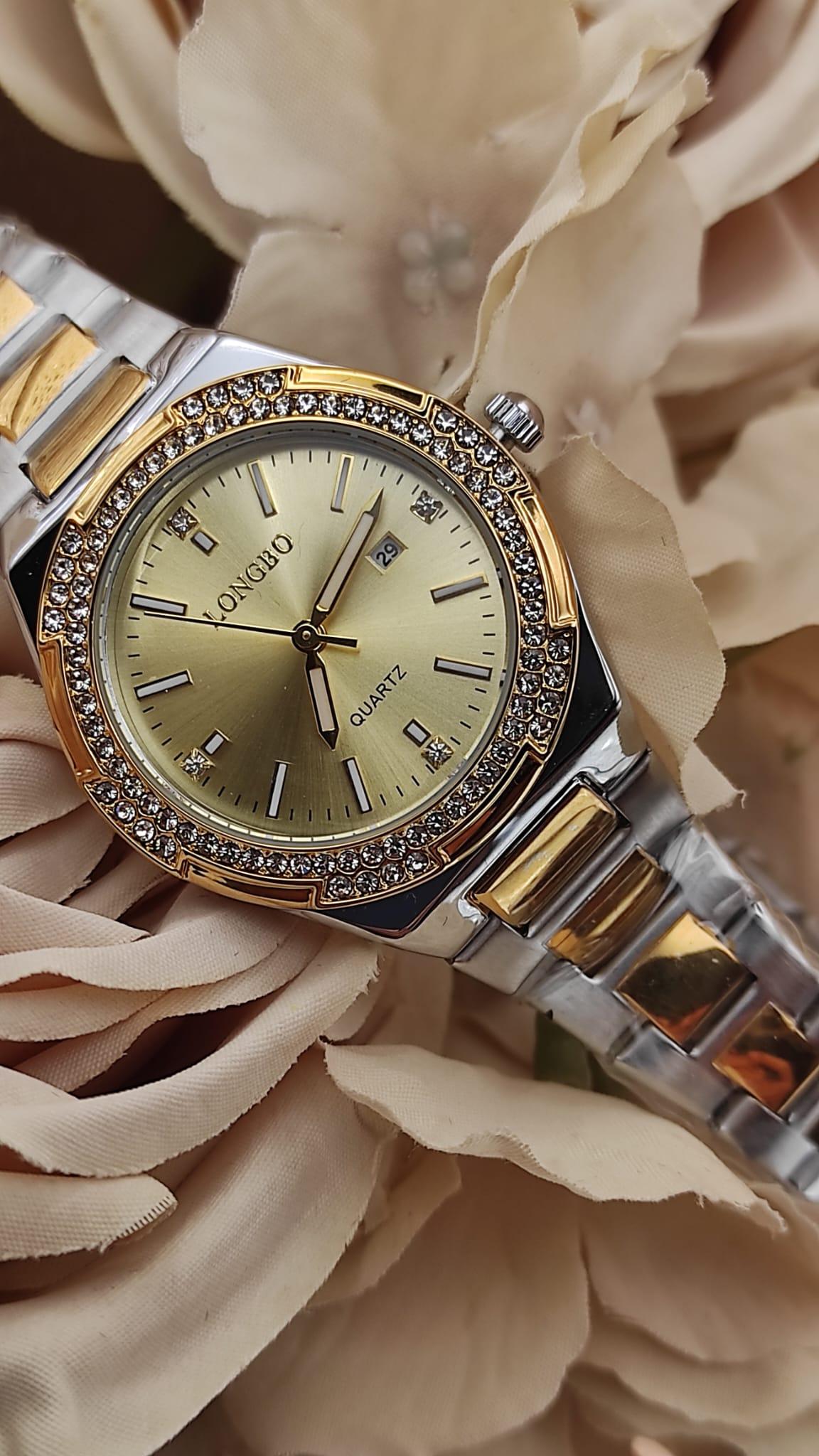Montre LONGBO à cadran doré avec bracelet bicolore argent et or, lunette sertie de cristaux brillants, posée sur un fond de fleurs beige.