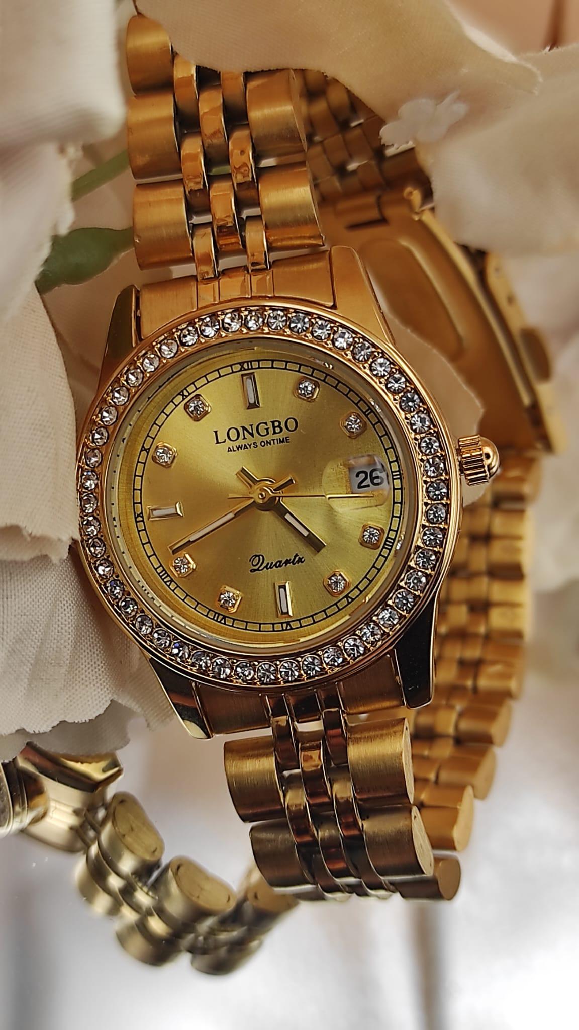 Montre LONGBO entièrement dorée avec cadran doré, lunette sertie de cristaux et bracelet métallique, posée sur un fond de pétales crème.