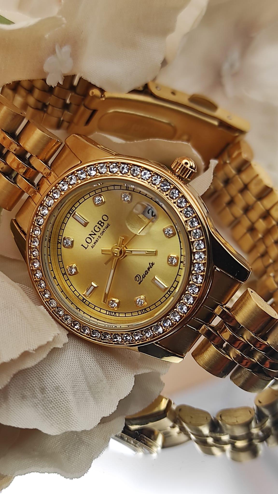 Montre LONGBO entièrement dorée avec cadran doré, lunette sertie de cristaux et bracelet métallique, posée sur un fond de pétales crème.