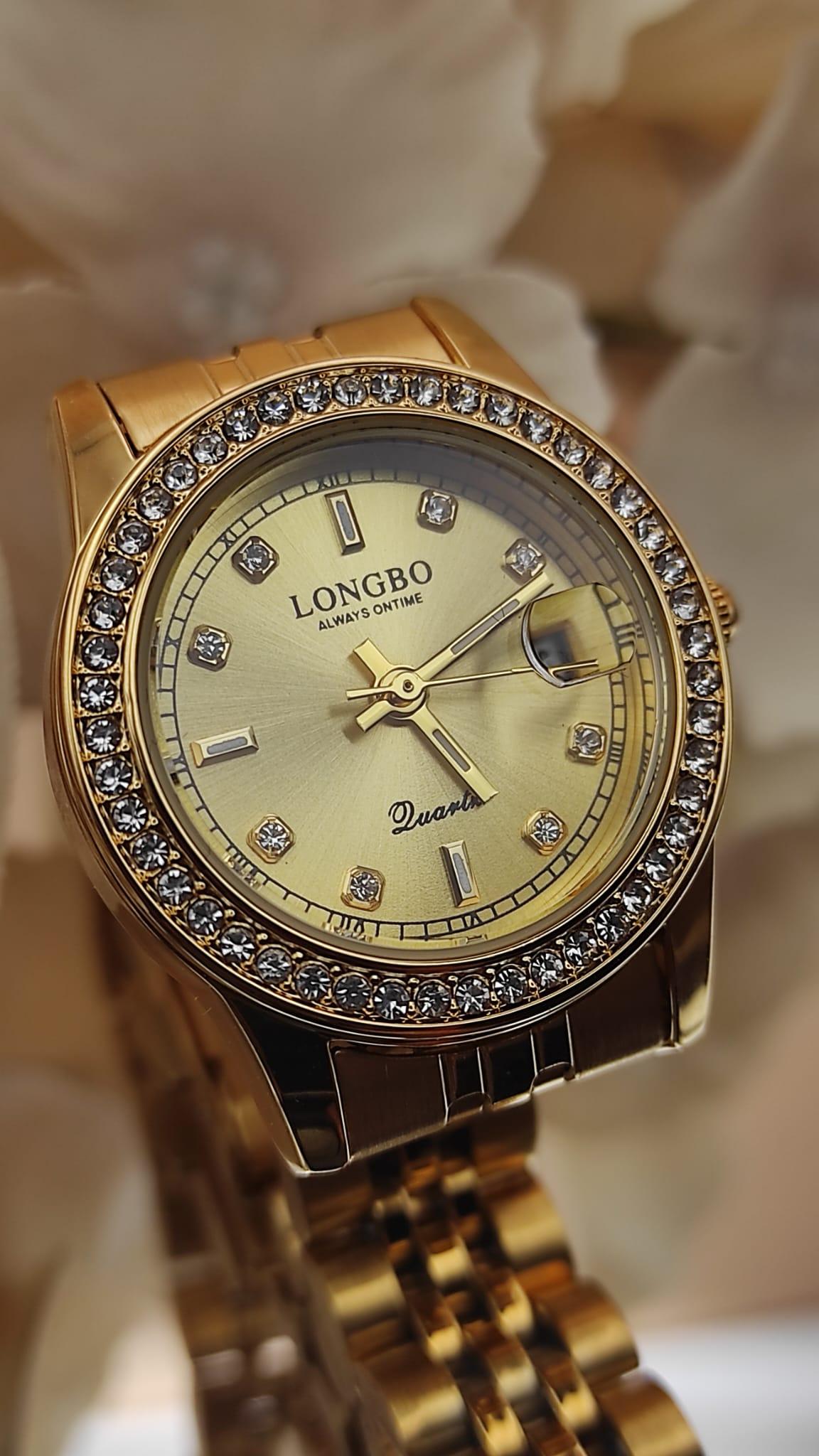 Montre LONGBO entièrement dorée avec cadran doré, lunette sertie de cristaux et bracelet métallique, posée sur un fond de pétales crème.