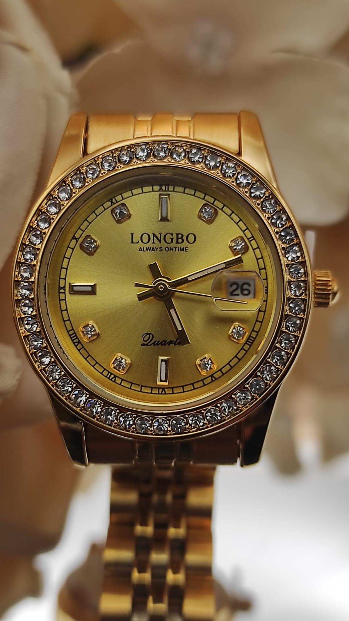 Montre LONGBO entièrement dorée avec cadran doré, lunette sertie de cristaux et bracelet métallique, posée sur un fond de pétales crème.