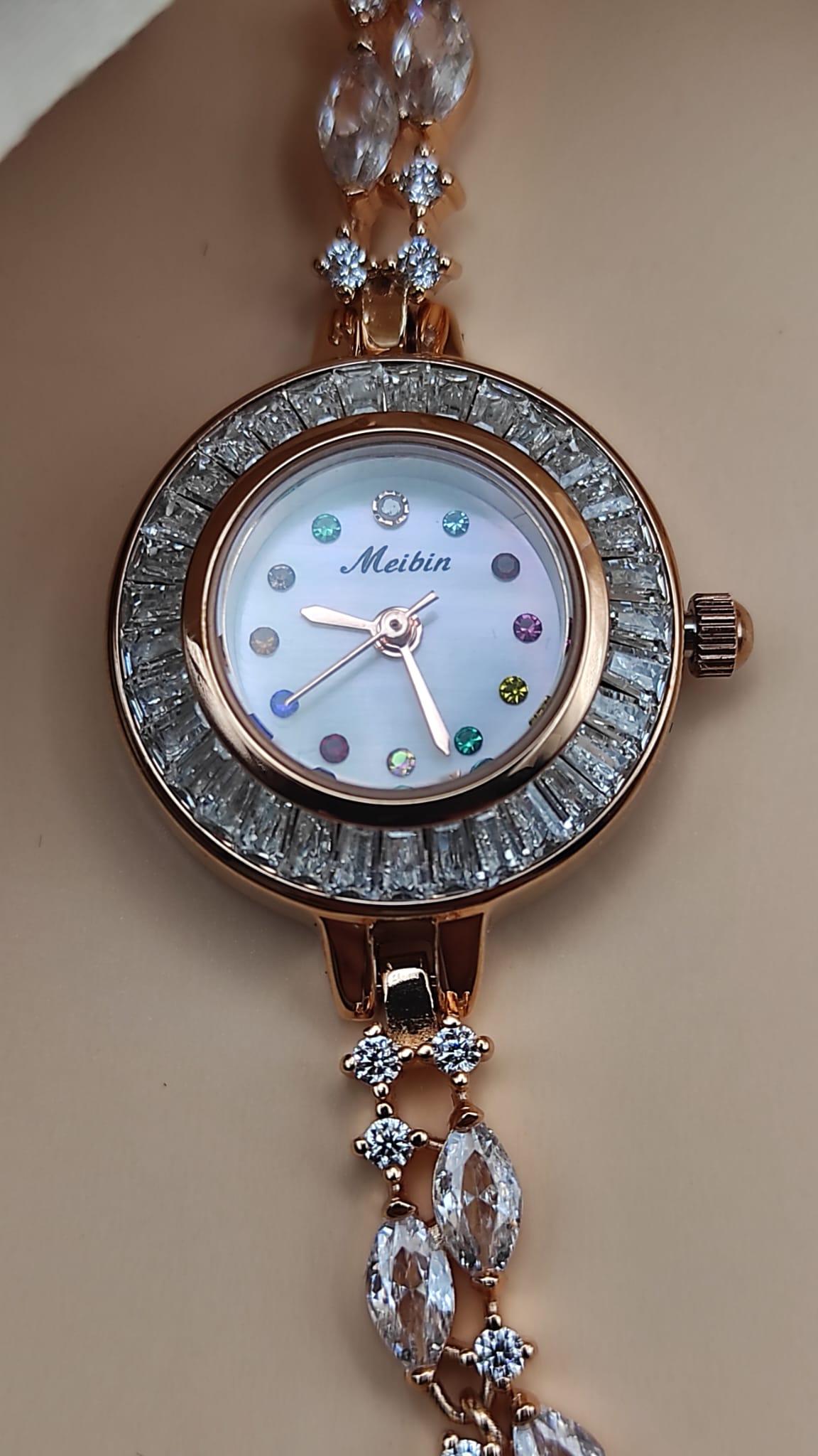 Montre Tima avec cadran blanc et index en pierres multicolores, lunette sertie de cristaux, bracelet rose gold orné de pierres décoratives, posée sur un fond de pétales beige.