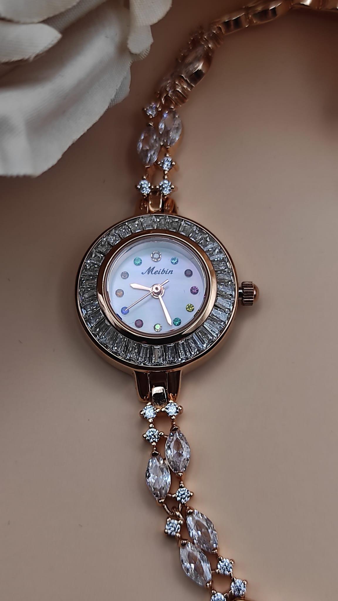 Montre Tima avec cadran blanc et index en pierres multicolores, lunette sertie de cristaux, bracelet rose gold orné de pierres décoratives, posée sur un fond de pétales beige.