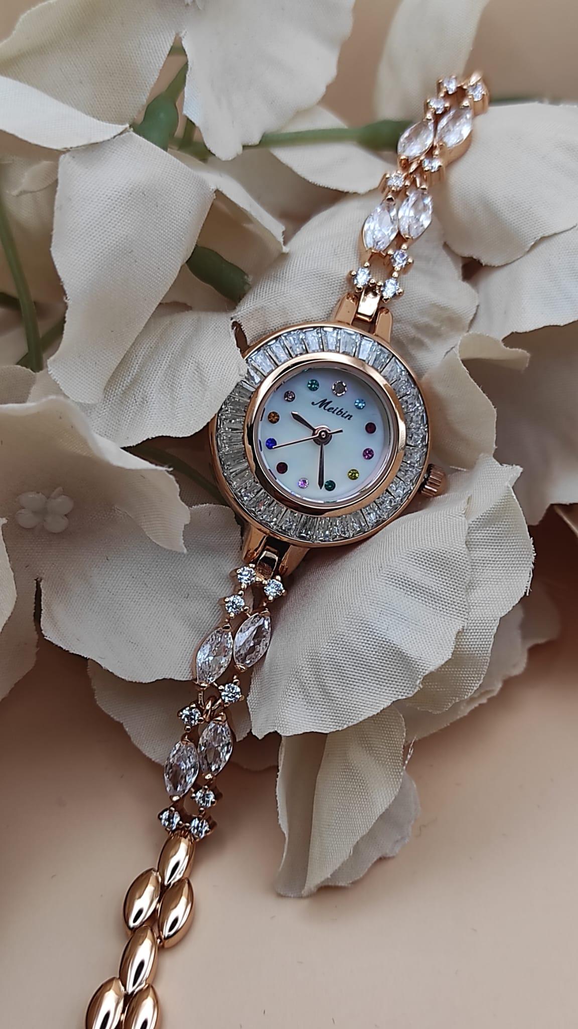 Montre Tima avec cadran blanc et index en pierres multicolores, lunette sertie de cristaux, bracelet rose gold orné de pierres décoratives, posée sur un fond de pétales beige.