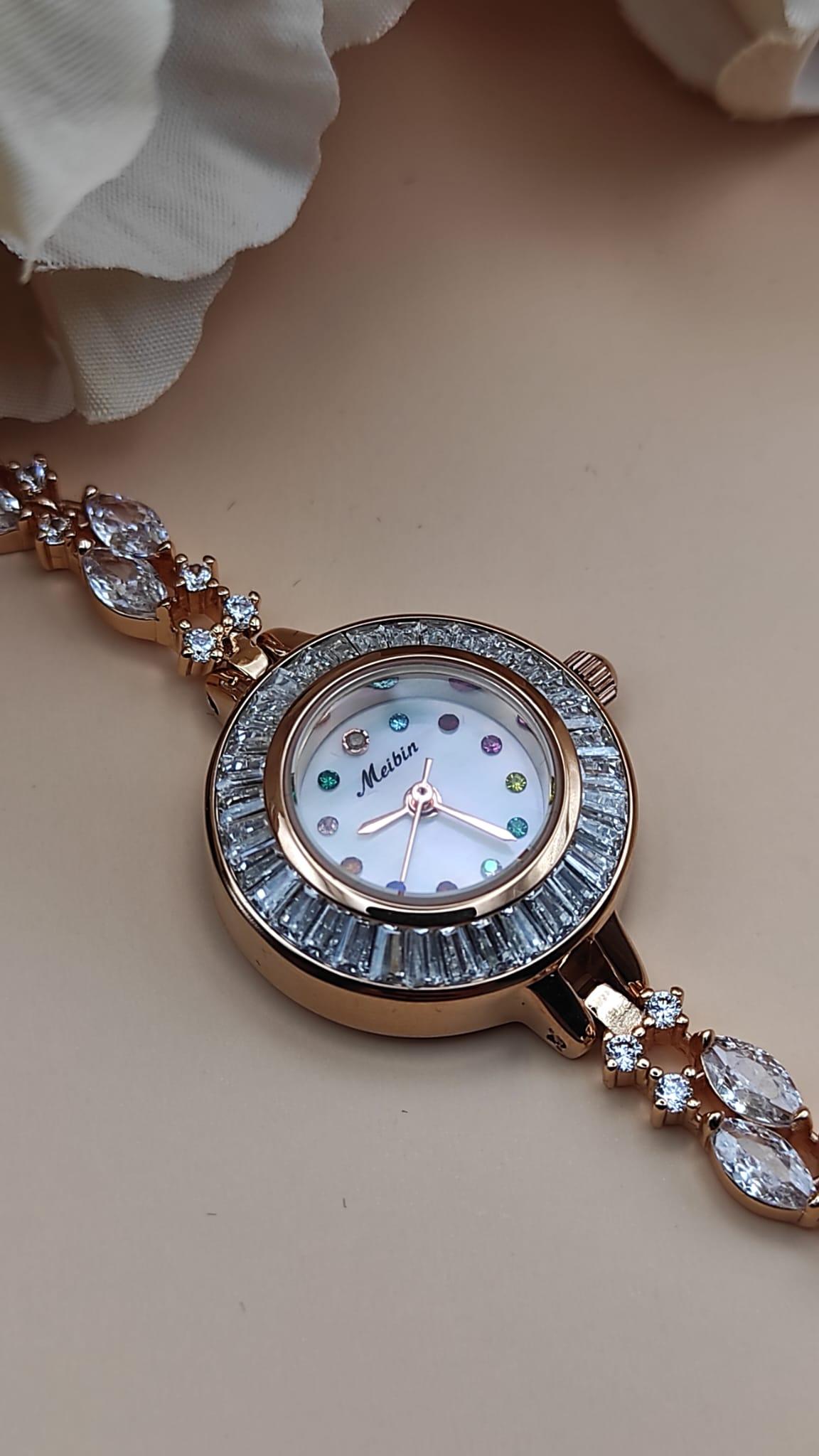 Montre Tima avec cadran blanc et index en pierres multicolores, lunette sertie de cristaux, bracelet rose gold orné de pierres décoratives, posée sur un fond de pétales beige.
