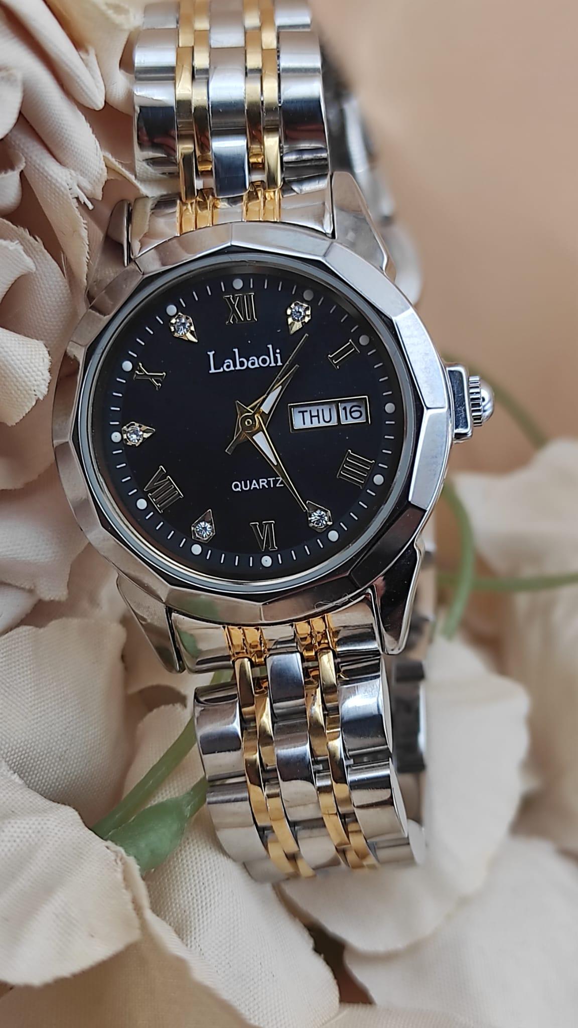 Montre Labaoli à cadran noir avec chiffres dorés stylisés, affichage jour et date à 3h, bracelet métallique bicolore argent et or, posée sur un fond floral beige.