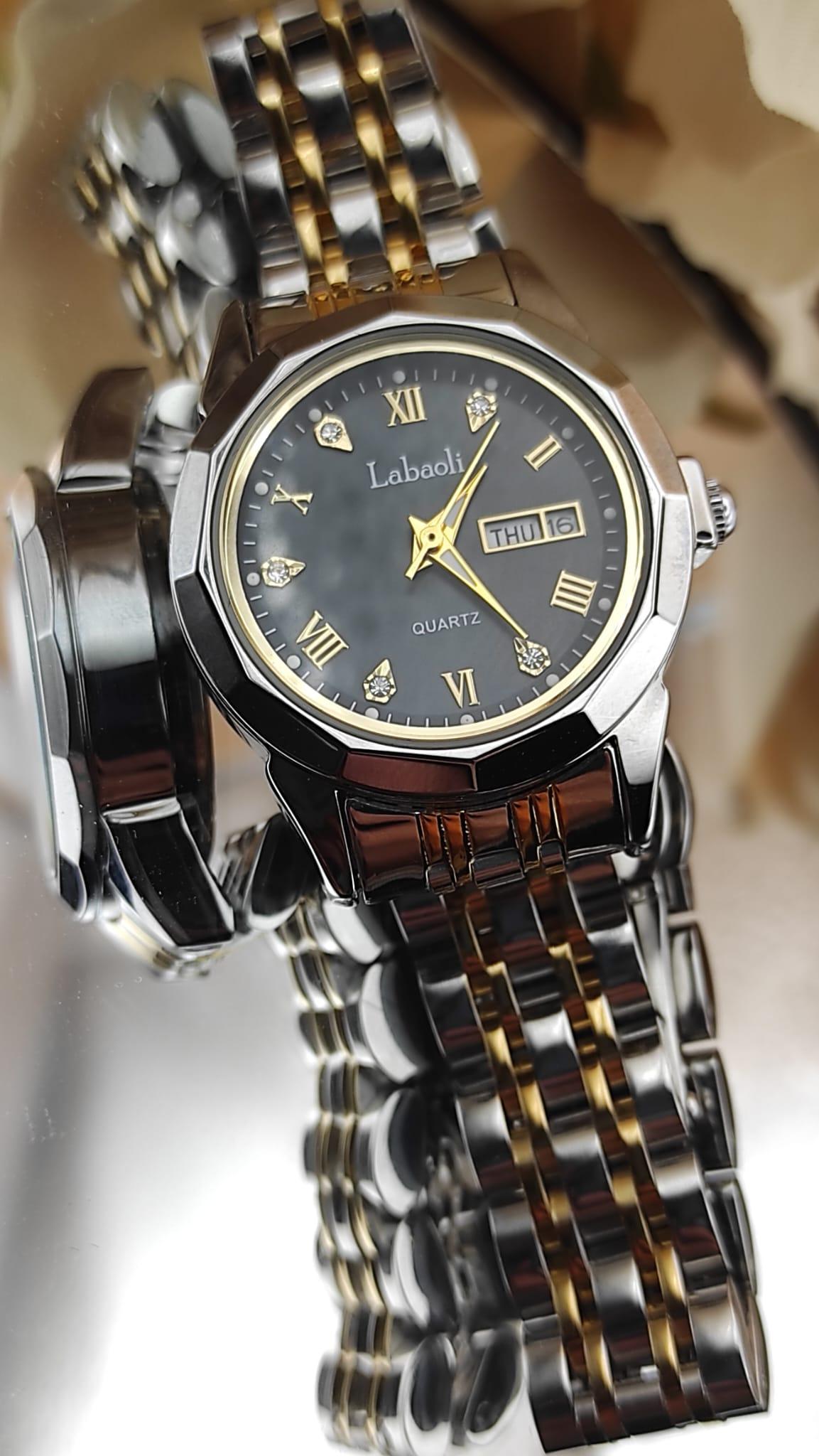 Montre Labaoli à cadran noir avec chiffres dorés stylisés, affichage jour et date à 3h, bracelet métallique bicolore argent et or, posée sur un fond floral beige.
