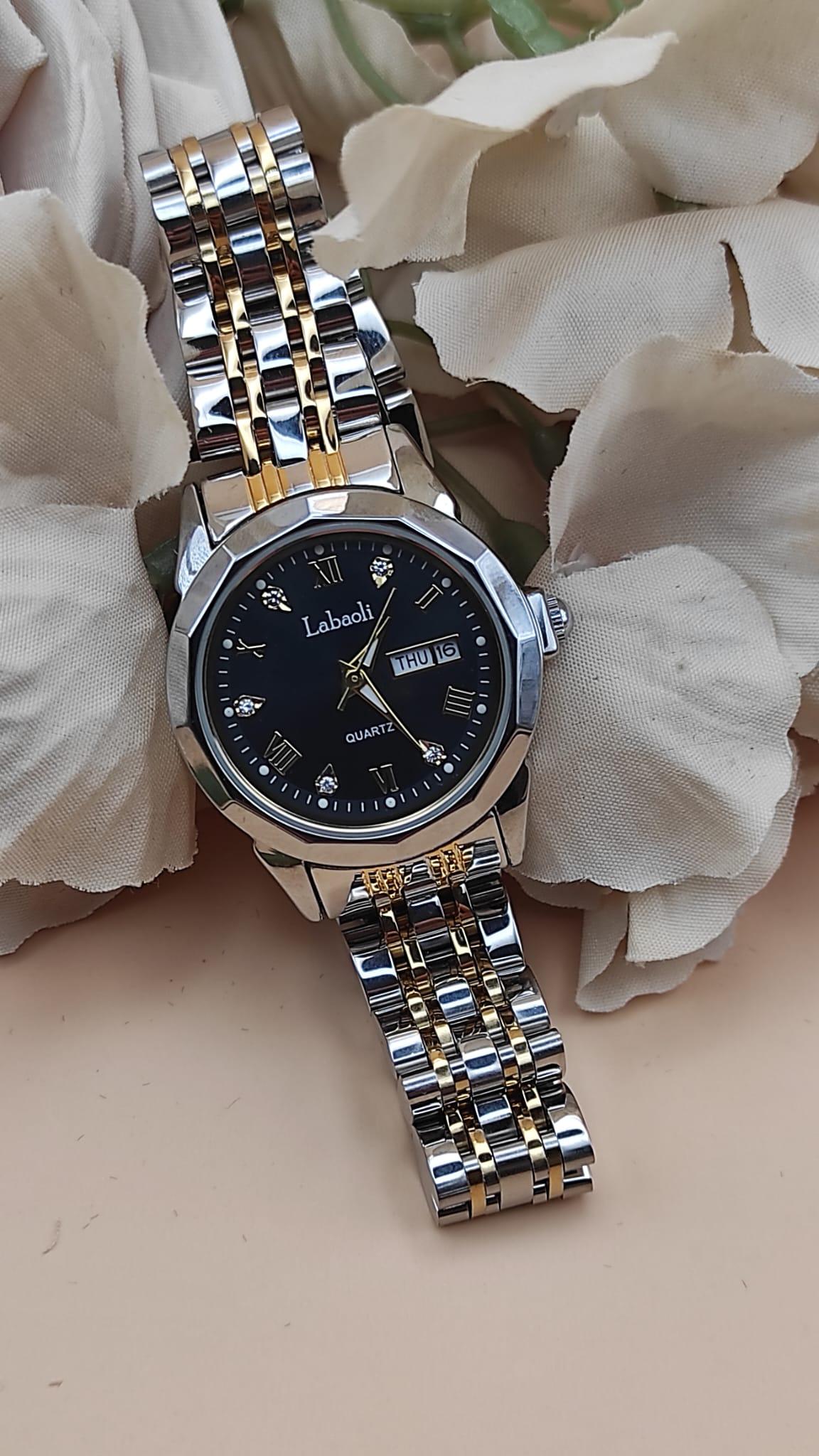 Montre Labaoli à cadran noir avec chiffres dorés stylisés, affichage jour et date à 3h, bracelet métallique bicolore argent et or, posée sur un fond floral beige.